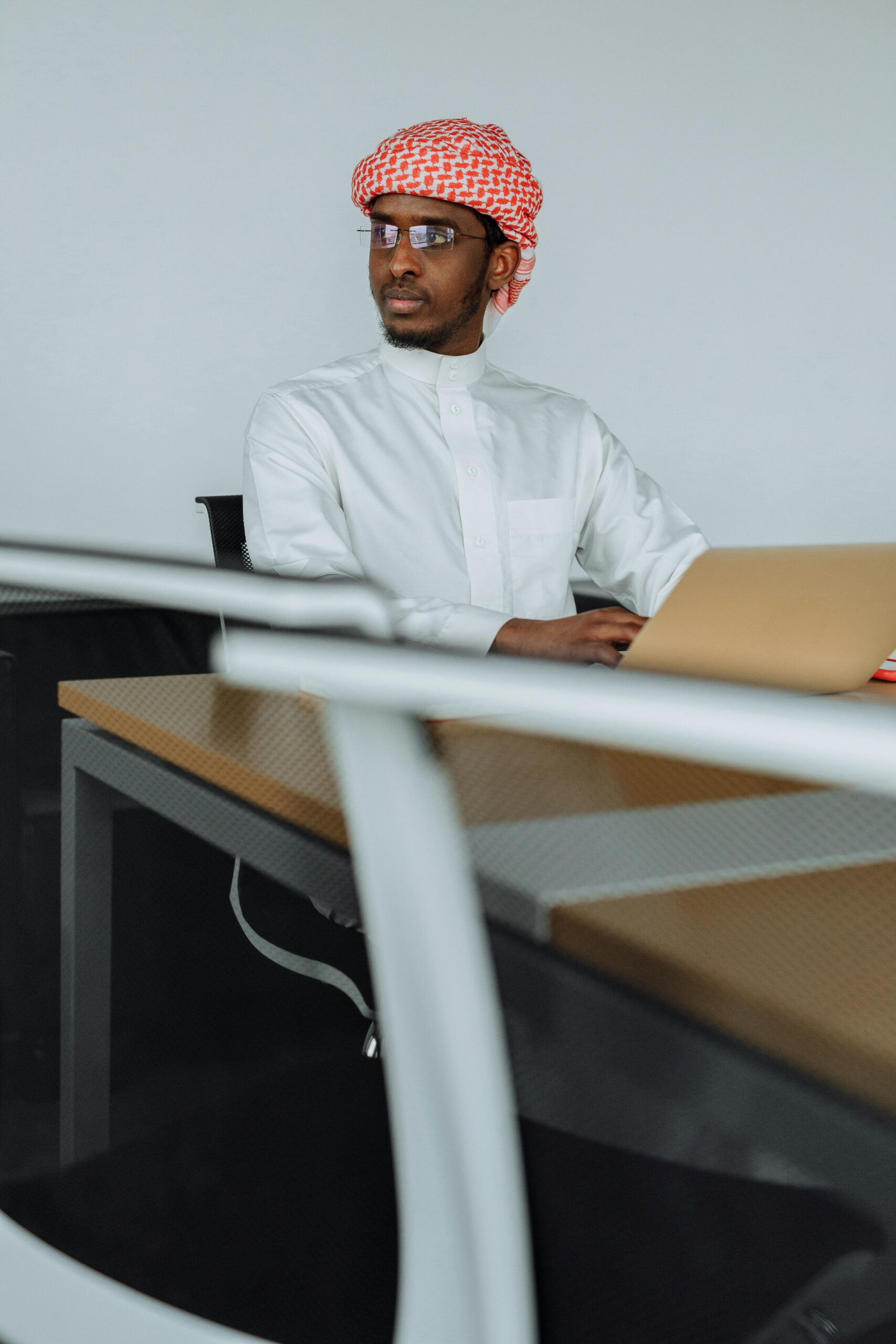 Home Middle Eastern man in traditional attire using laptop in modern office setting.